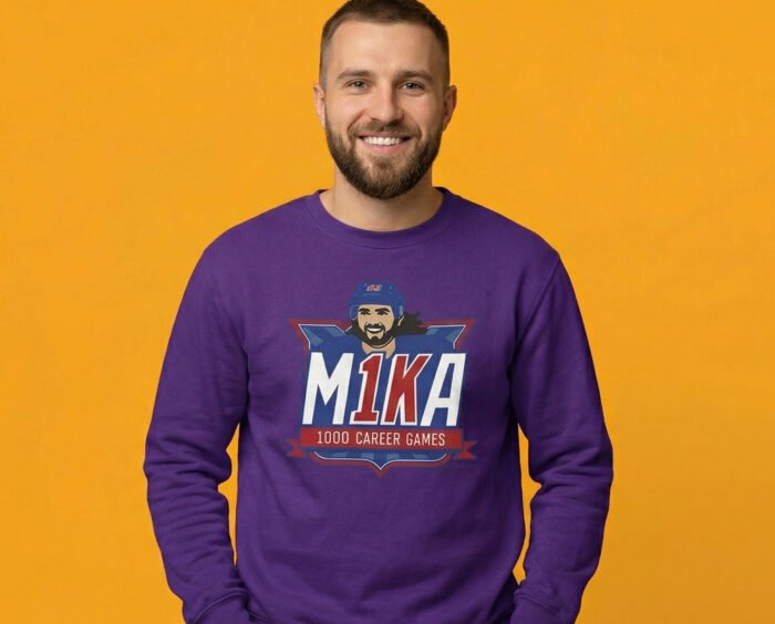 A smiling man wearing a purple long-sleeve sweatshirt with the 'M1KA 1000 Career Games' hockey milestone logo printed on the chest. The background is a vibrant mustard yellow."